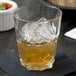 A stackable Arcoroc tall rocks glass with ice and amber liquid on a table with a bowl of food.