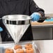 A person pouring powder through a Vollrath Coarse China Cap Strainer onto doughnuts.