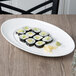 A white 10 Strawberry Street oval porcelain platter with sushi rolls on a wood table.