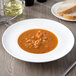 A 10 Strawberry Street white porcelain soup bowl with soup on a table with a slice of bread and a knife.