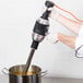 A person using a KitchenAid immersion blender to mix liquid in a metal container.