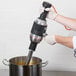 A person using a KitchenAid blending arm to mix soup in a metal container.