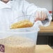 A person in a white coat using a Cambro clear polycarbonate scoop to serve macaroni from a plastic container.