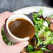 A person using a Chef & Sommelier Geode stoneware bowl to pour sauce over a salad.