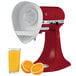 A KitchenAid citrus juicer attachment juicing oranges with a red KitchenAid mixer in the background.