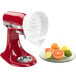 A white KitchenAid citrus juicer attached to a red KitchenAid mixer with a plate of citrus fruit.