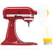 A red KitchenAid mixer with a KitchenAid citrus juicer attachment juicing oranges into a glass.