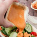 A hand holding a paper bowl of salad with a sauce being dipped into.