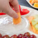 A person dipping a carrot into a bowl of fruit using an EcoChoice palm leaf deli tray with fruit in a compartment.