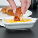 A person holding a hot dog and dipping it into a Tuxton bright white rectangle china sauce dish full of mustard.