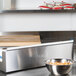 A silver metal Edlund Film Dispenser Blade in a school kitchen on a counter above a bowl of noodles.