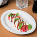 A Libbey ivory porcelain platter with sliced tomatoes, basil, and olive oil on a table.