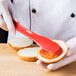 A person using an HS Inc. red polypropylene sandwich spreader to spread jam on a piece of bread.