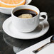 A Libbey white porcelain tea saucer with a cup of coffee on a table with a spoon and orange slices.