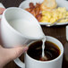 A hand pouring milk into a white Libbey porcelain creamer over a white cup of coffee.