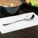 A Libbey stainless steel bouillon spoon on a napkin next to a bowl of soup.