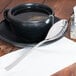 A Libbey stainless steel teaspoon on a table next to a cup of tea.