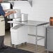 A man in a chef's uniform standing in front of a Regency stainless steel open base work table.