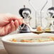 A hand holding a Libbey stainless steel round soup spoon over a bowl of soup with a meatball on it.