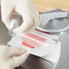 A gloved hand uses a machine to cut raw meat on a metal counter with a Garde Hamburger Patty Paper tube.