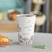 A close-up of a Choice double wall paper hot cup filled with coffee on a table with a croissant.