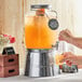 A person pouring orange juice into an Acopa Mason Jar Glass Beverage Dispenser on a table in an outdoor catering setup.