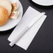 A Libbey stainless steel bread and butter knife on a napkin next to a plate of bread.