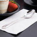 A Libbey stainless steel dessert spoon on a napkin next to a plate of cake.