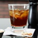 An Anchor Hocking Omega old fashioned glass filled with brown liquid, ice, and nuts on a table.