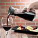 A person pouring red wine from an Arcoroc Fluid Carafe on a table with a plate of fruit and cheese.