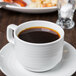 A Tuxton Pacifica white china cup filled with coffee on a saucer on a white table.