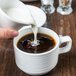 A person pouring milk into a Tuxton Pacifica china cup of coffee.