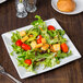 A plate of salad with tomatoes, lettuce, and croutons on a Tuxton bright white square china plate.