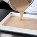 A person using a bowl to pour brown liquid into a white fiberglass sheet pan with a pan extender.