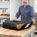 A woman in a chef's hat holding a black ServIt insulated pizza delivery bag on a counter with a pizza box inside.