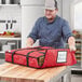 A man in a blue shirt and cap looking at a red CaterGator Dash insulated pizza bag with pizza boxes inside.