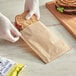 A person in gloves putting a sandwich in a Kraft paper bag.