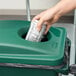 A hand putting a paper into a green Rubbermaid Slim Jim recycling container.