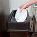 A hand holding a white cup in a Continental black wall hugger trash can.