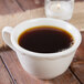 A close up of a Tuxton eggshell white china cup filled with coffee on a table with a lit candle.