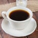 A Tuxton eggshell white china saucer with a cup of coffee on it.