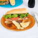 A sandwich with lettuce and tomatoes on a Creative Converting pumpkin spice orange paper plate with a knife and fork next to it.