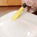 A hand using a yellow and black Mercer Culinary saw tooth silicone brush to plate a white dish.
