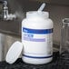 A white container with a lid of bar glass cleaner powder with a plastic spoon inside.