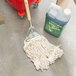 A white Rubbermaid wet mop in a red bucket next to a gallon of detergent.