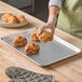 A half size aluminum perforated bun or sheet pan with croissants being placed on it.