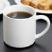 A white Libbey porcelain mug filled with brown liquid sits on a table next to a plate of food.