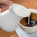 A hand pouring milk from a white Libbey porcelain creamer into a cup of coffee.