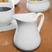 A close-up of a white Libbey porcelain creamer on a wood table with a cup of coffee.