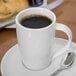 A Libbey Royal Rideau white porcelain mug filled with coffee on a plate with a spoon and pastry.